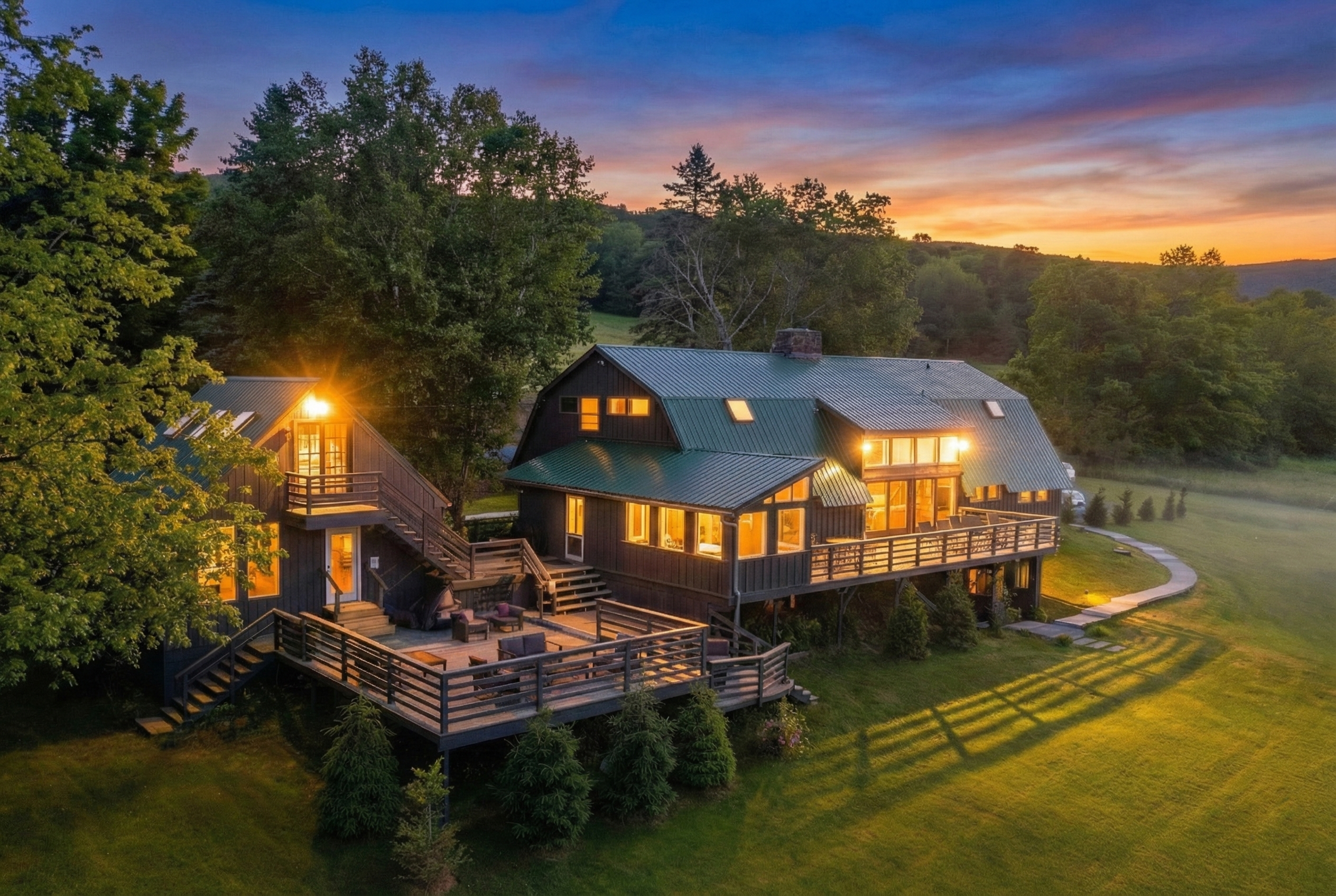 Spacious, well-lit living room and dining area with large windows and glass sliding doors on the left, leading to an outdoor deck with chairs. Inside, there are brown armchairs, a white sofa, a wooden coffee table with plants, and a white bench. A stone fireplace is on the right, and a wooden dining table with chairs is in the background. Ceiling fans and potted plants are also visible.