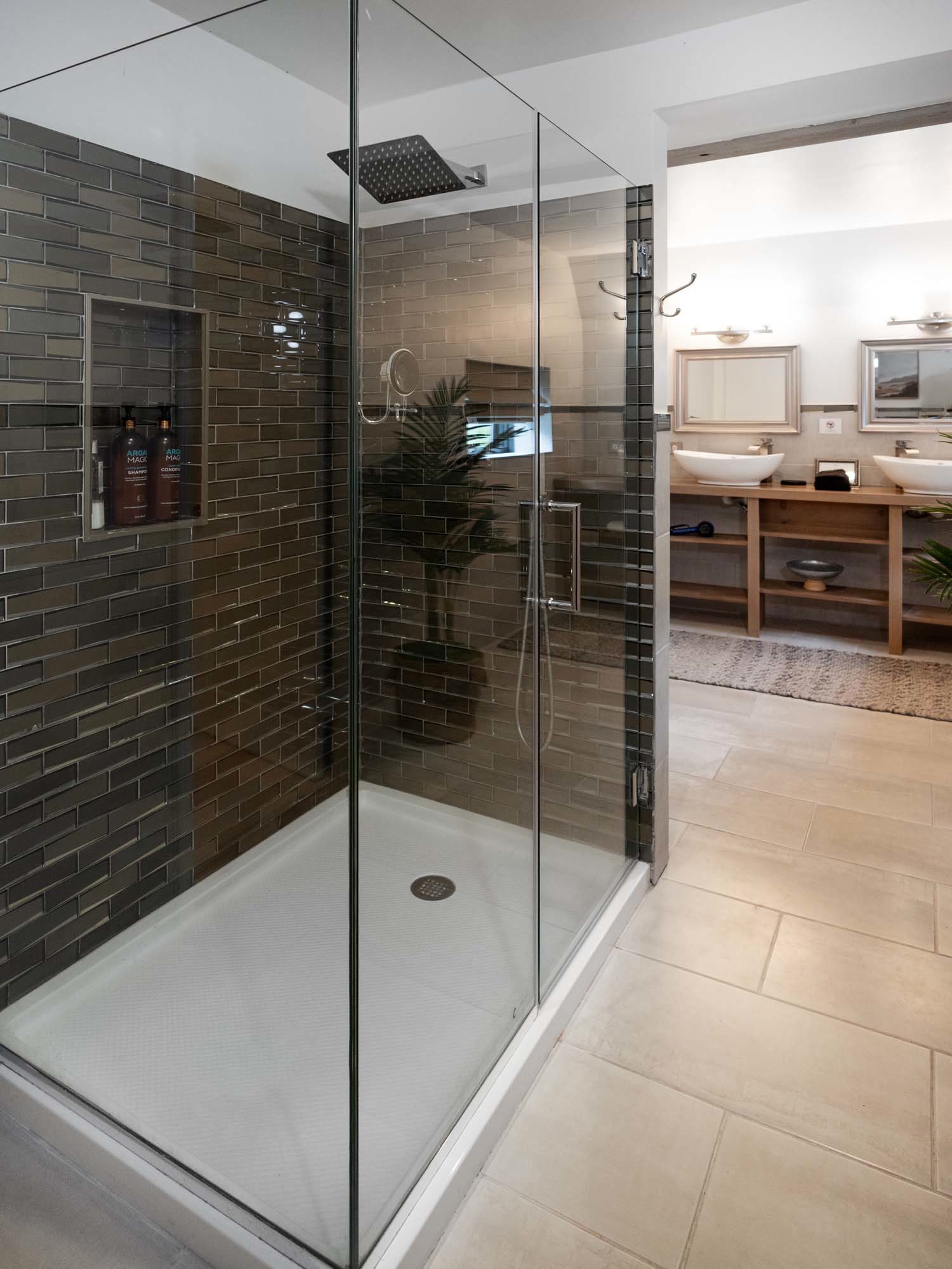 Modern bathroom with a glass-enclosed shower area featuring dark brown rectangular tiles on the walls. Inside the shower, there are two bottles labeled shampoo and conditioner on a built-in shelf. The shower has a square overhead rain showerhead and a handheld showerhead. Outside the shower, the bathroom floor is tiled in light beige. In the background, there is a double sink vanity with two vessel sinks, two framed mirrors, and wall-mounted lights above each mirror.