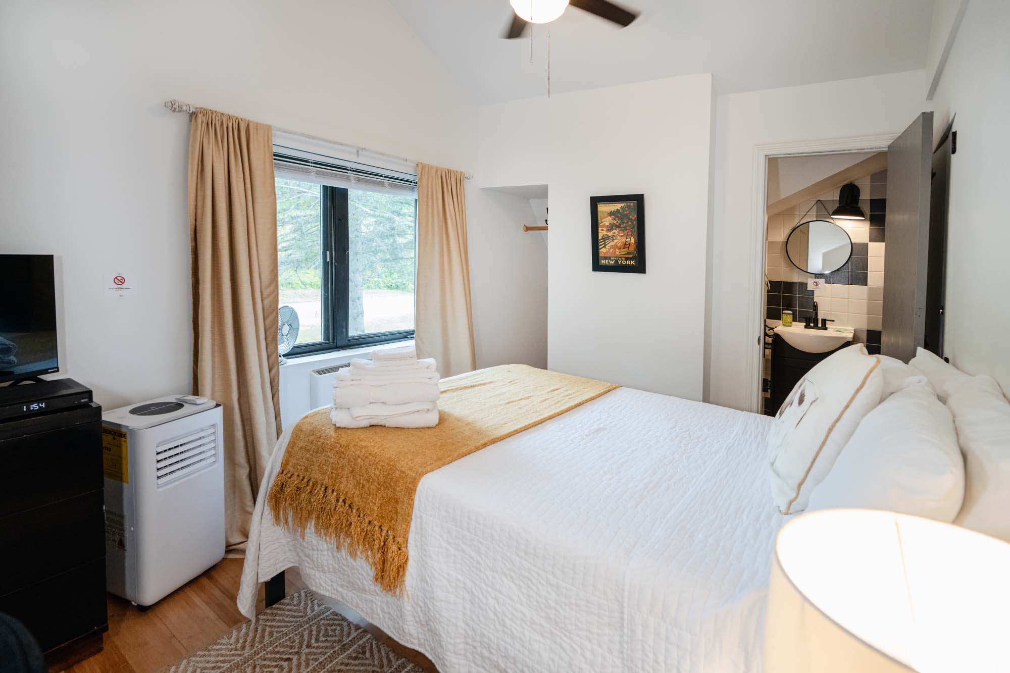 Bedroom with a neatly made bed covered in white bedding and a mustard-colored throw blanket. A stack of white towels is placed on the bed. The room has beige curtains covering a window. To the left, there is a black dresser with a TV on top and a white portable air conditioner beside it. A framed picture hangs on the wall. An open door reveals a bathroom with a round mirror and a sink.