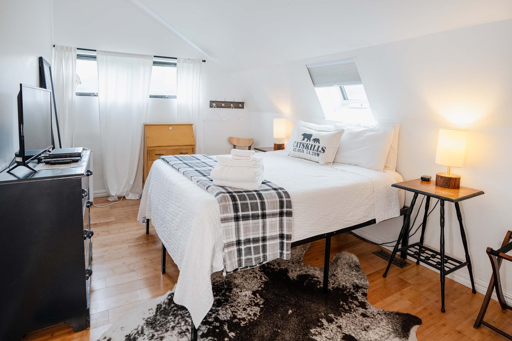 Bright bedroom with white walls and wooden flooring. A bed with white bedding and a gray plaid runner is centered, topped with folded white towels and a pillow labeled ‘CATSKILLS.’ On each side of the bed are small wooden tables with lit lamps. A black dresser with a TV and electronic devices sits to the left. A wooden chair and a small wooden cabinet are near the window. A black and white rug is partially visible under the bed.