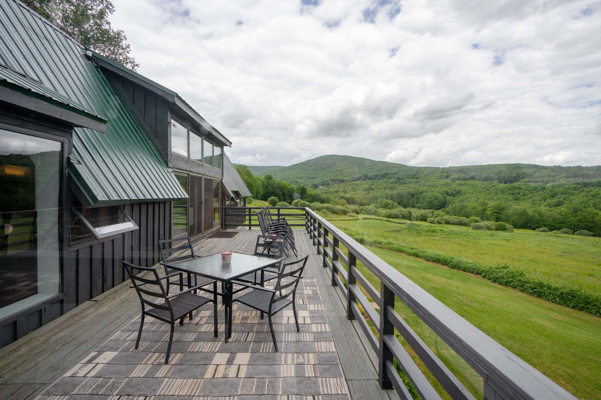 Wooden deck attached to a dark-colored building with a green metal roof. On the deck, there is a glass-top table with four metal chairs surrounding it, and a small cup on the table. Further along the deck, there are several black chairs lined up against the railing. The deck overlooks a large green field with trees and hills in the background under a cloudy sky.