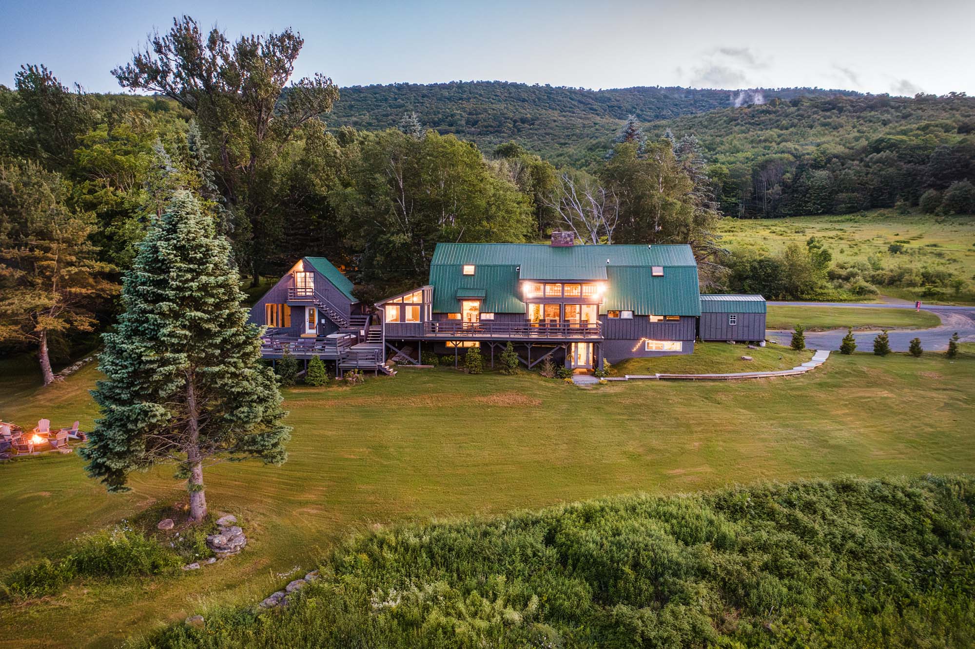 Large house with a green metal roof and dark exterior walls situated in a grassy clearing surrounded by trees. The house has multiple sections, including a main building and an adjacent smaller structure with an external staircase. Several windows are illuminated, casting warm light. In the foreground, there are two tall evergreen trees and a fire pit area with chairs arranged around it. A paved pathway and driveway are visible on the right side, with a forested hill in the background.