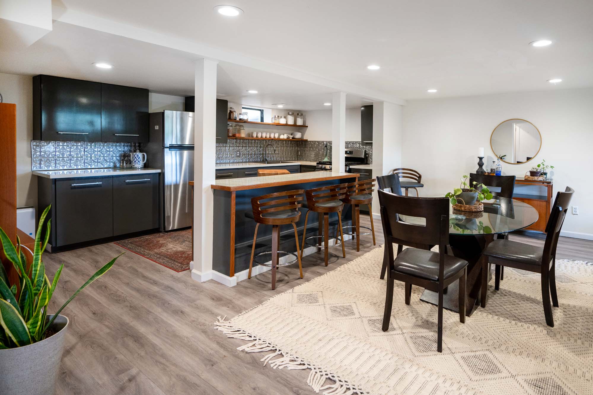 Modern kitchen and dining area with light wood flooring and white walls. The kitchen has dark cabinets, a stainless steel refrigerator, a stove, and a patterned backsplash. A wooden breakfast bar with three wooden stools is positioned in front of the kitchen. The dining area features a round glass table with six dark wooden chairs on a large beige patterned rug. There is a round mirror and a small side table with plants and decor on the right side.