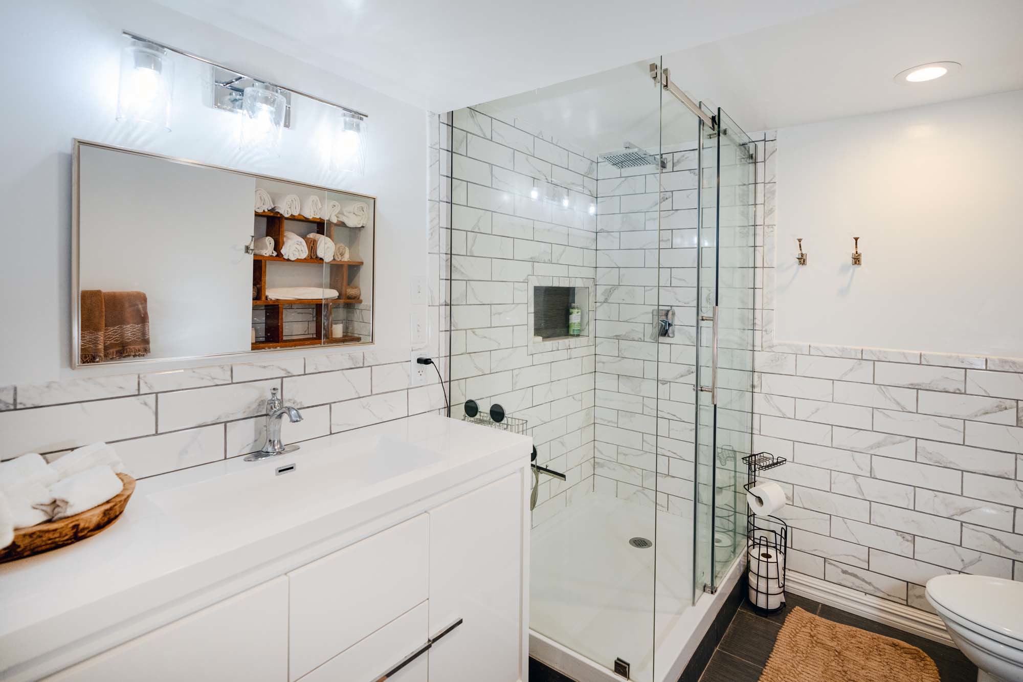 Modern bathroom with white subway tiles on the walls and dark floor tiles. There is a glass-enclosed shower with a built-in shelf and a rain showerhead. A white vanity with a sink and a chrome faucet is on the left side, topped with a basket of rolled towels. Above the vanity is a mirror with a wooden shelf unit inside, holding more rolled towels. On the right, there are two hooks on the wall, a toilet paper holder, and a beige bath mat on the floor.