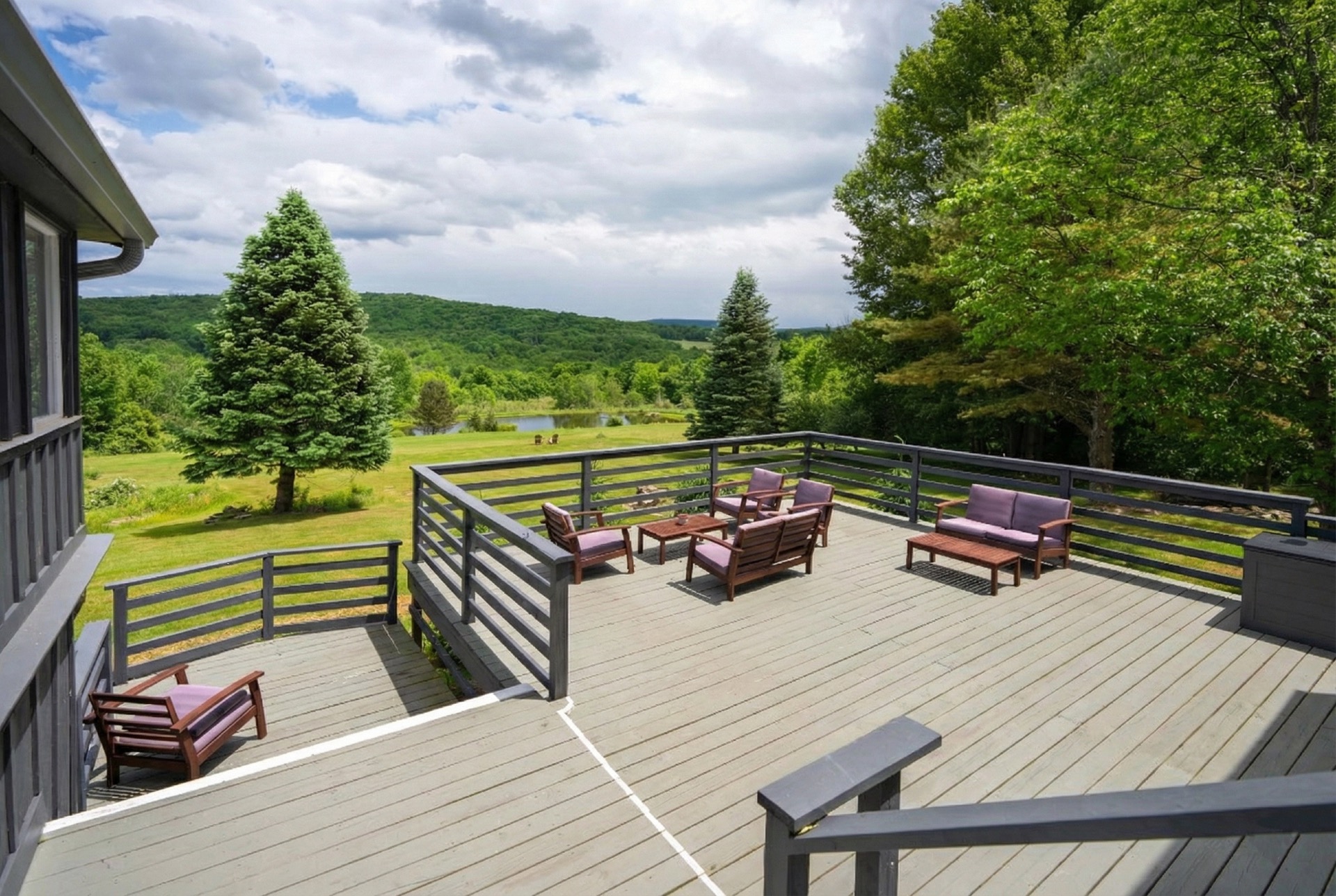 Spacious wooden deck with gray railings and flooring. The deck has outdoor furniture including two armchairs, a loveseat, and a small table, all with purple cushions. There is a staircase leading down from the deck. Surrounding the deck is a grassy area with trees, including two prominent evergreen trees and a pond in the background under a partly cloudy sky.