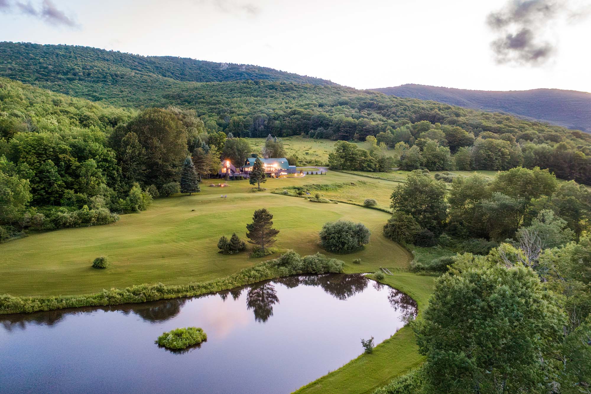 Serene landscape with a small pond in the foreground, surrounded by green grass and bushes. In the middle distance, there is a house with lights on, nestled among trees. The background features densely forested hills and mountains under a sky with some clouds. The scene is peaceful and natural.