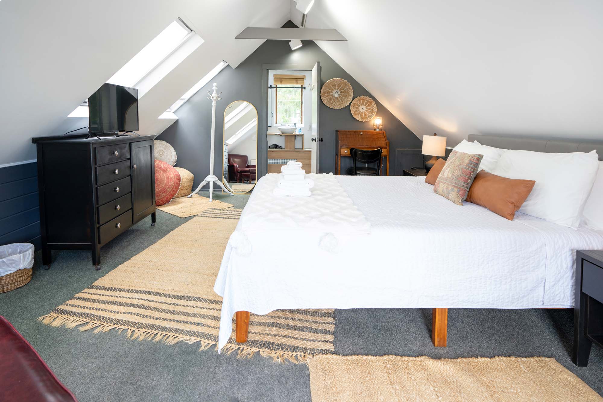 Bedroom with a sloped ceiling and two skylights. A large bed with white bedding and multiple pillows is centered, with folded towels on top. To the left is a black dresser with a TV on it. A full-length mirror, a white coat rack, and woven baskets are near the back wall. A small desk with a chair and a lamp is on the right side. The floor is carpeted with multiple rugs.