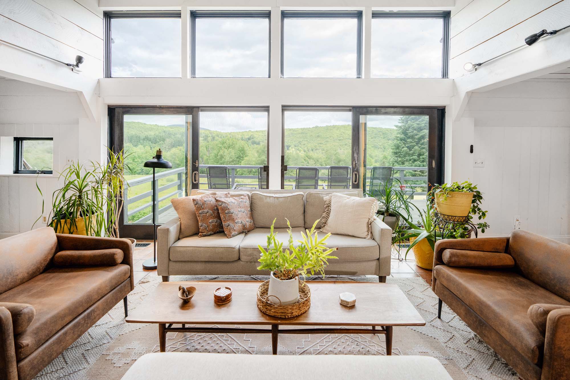 Living room with large windows and sliding glass doors revealing a green, forested landscape outside. Inside, there is a beige sofa with patterned and plain pillows, flanked by two brown leather armchairs. A wooden coffee table in front of the sofa holds a potted plant and small decorative items. Several potted plants are placed near the windows. The room has white walls and a light-colored rug with geometric patterns.