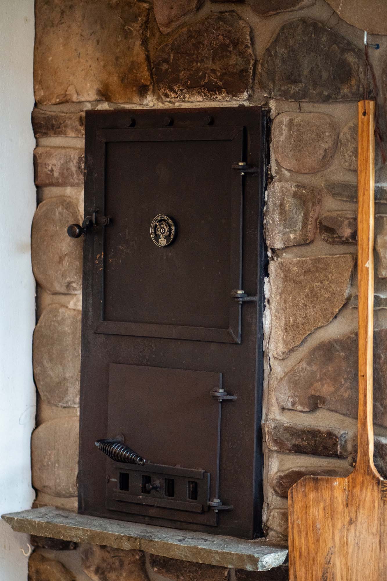 Black metal oven or furnace door set into a stone wall. The door has two sections: a larger upper door with a round temperature gauge or dial in the center, and a smaller lower door with a spring handle and vent openings. To the right of the door, there is a wooden paddle or peel leaning against the stone wall.