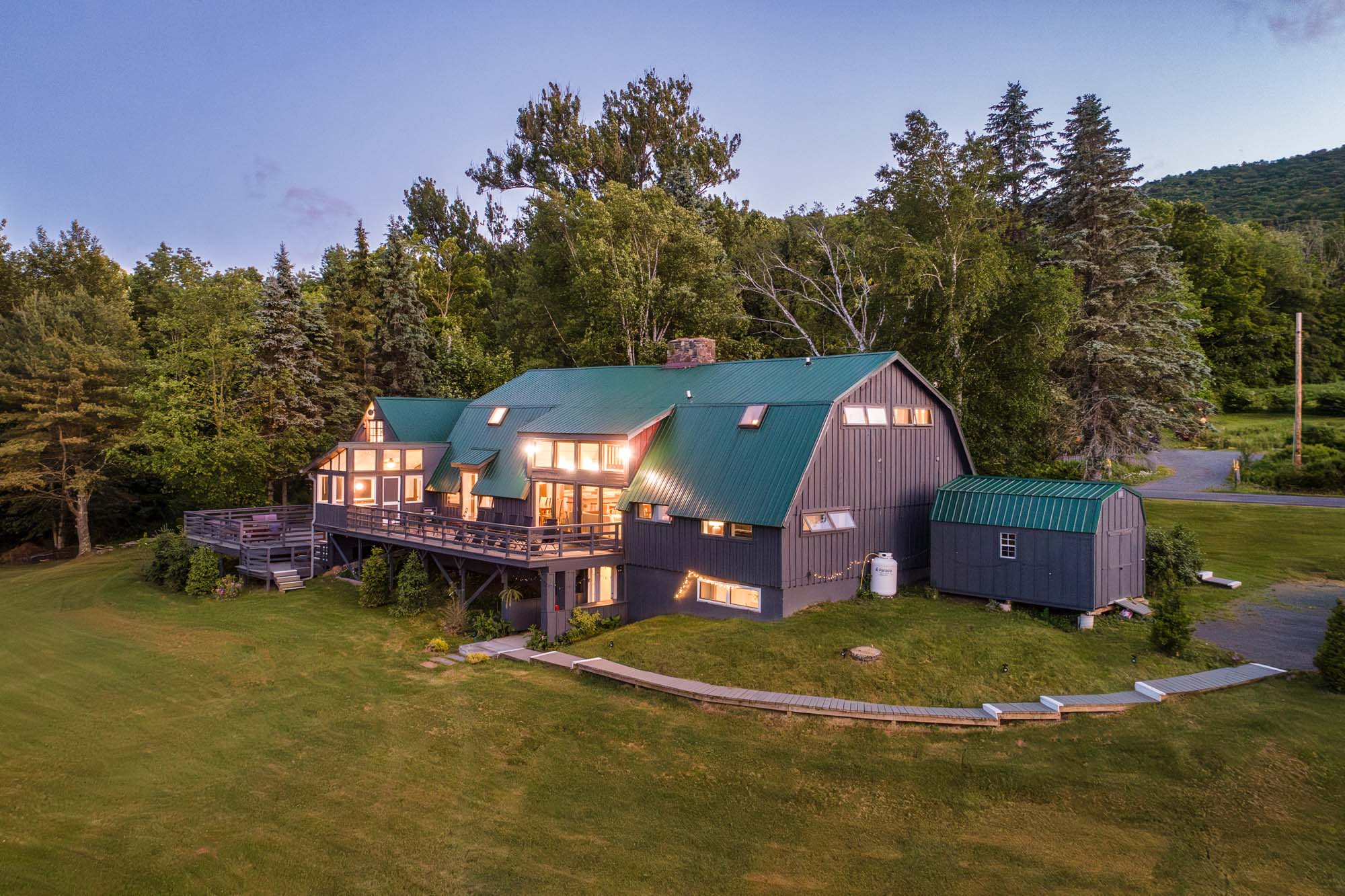 Large house with a green roof and dark gray walls situated on a grassy lawn. The house has multiple windows, some of which are lit from the inside. There is a wooden deck attached to the house and a small detached shed with a matching green roof nearby. The background features dense trees and a clear sky at dusk.