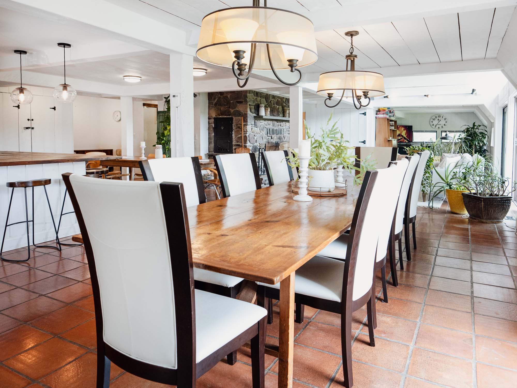 Spacious dining area with a long wooden table surrounded by eight chairs with white upholstery and dark wooden frames. Above the table, there are two large ceiling light fixtures with white lampshades. The floor is covered with terracotta tiles. In the background, there is a stone fireplace, a kitchen island with bar stools, and several plants near a window. The room has white walls and ceiling with exposed beams.